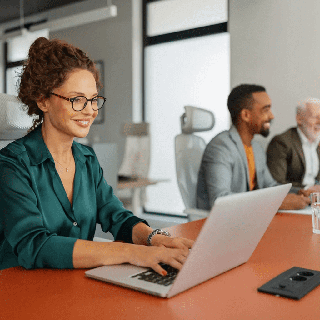 Smiling woman working on a laptop in a modern office with colleagues.